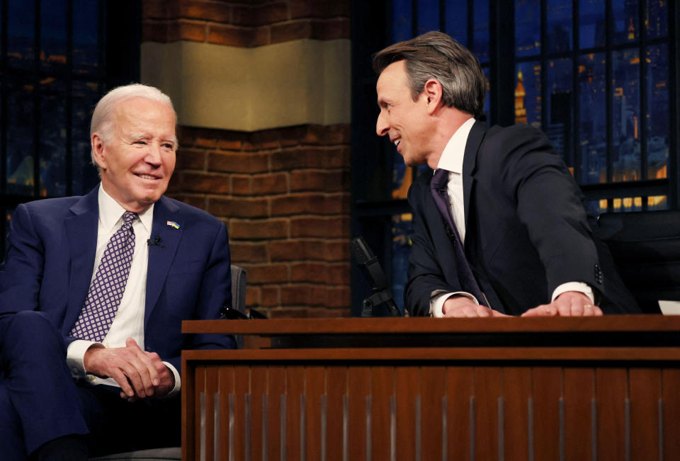 Biden seated in a chair next to Seth Meyers, seated behind a wooden desk.