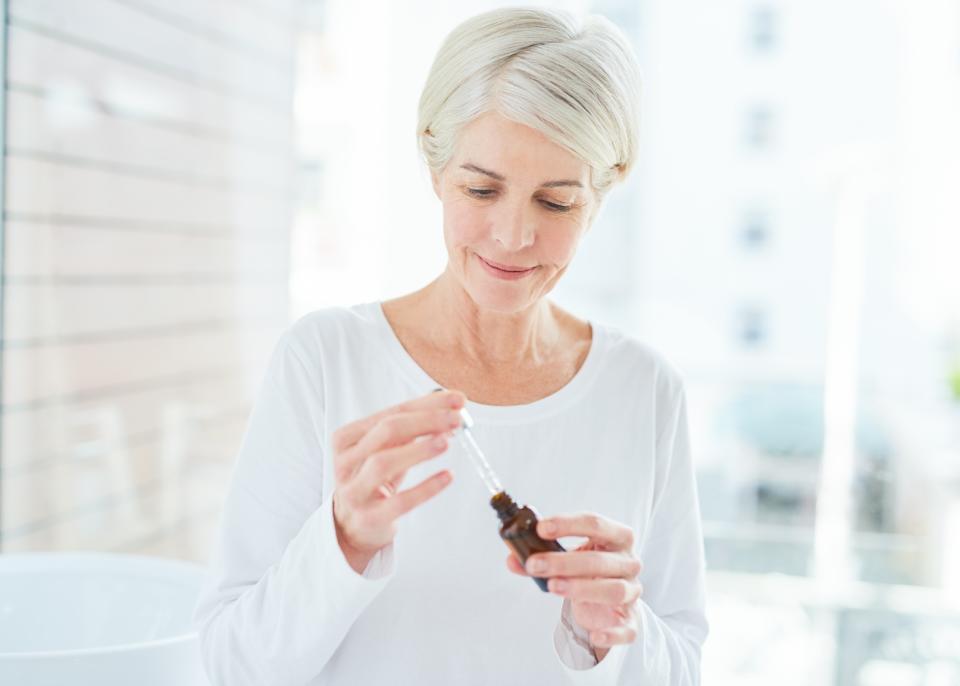 Rose Essential Oil Benefits: Shot of a mature woman holding a bottle of serum in a bathroom at home