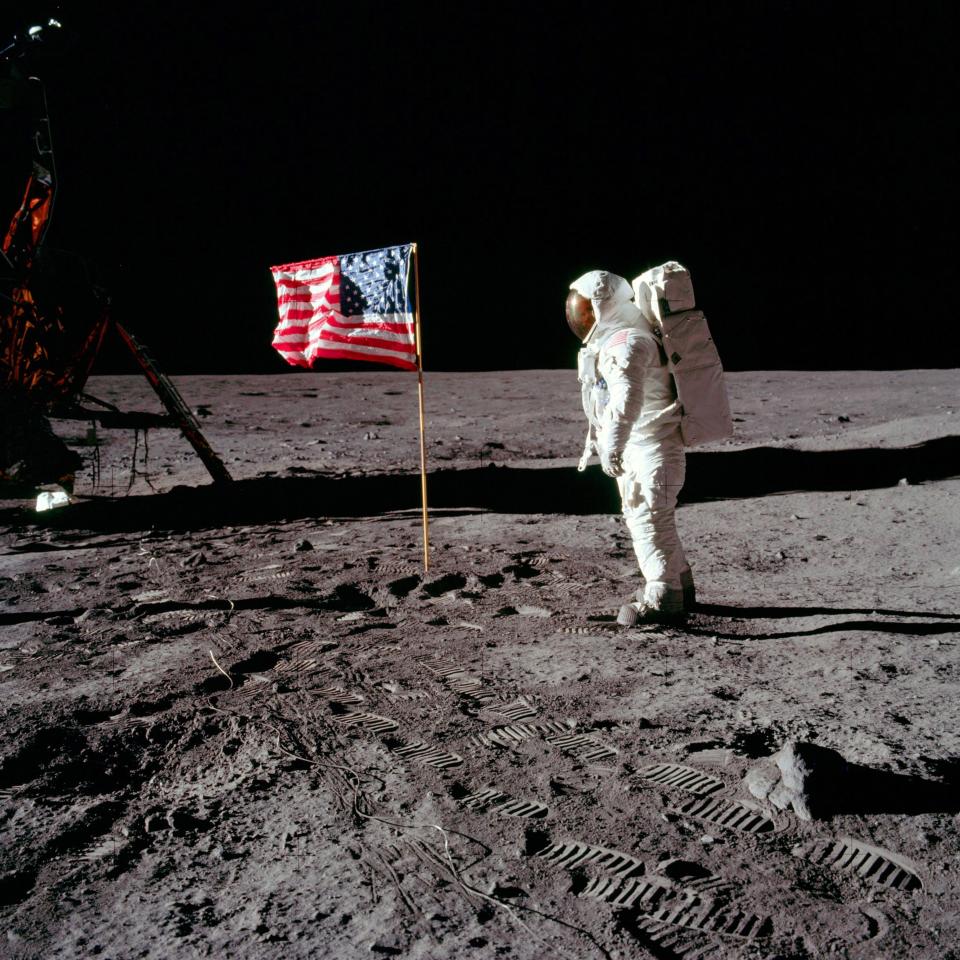 Astronaut Neil Armstrong stands on the moon next to the American flag with the lunar module in the background