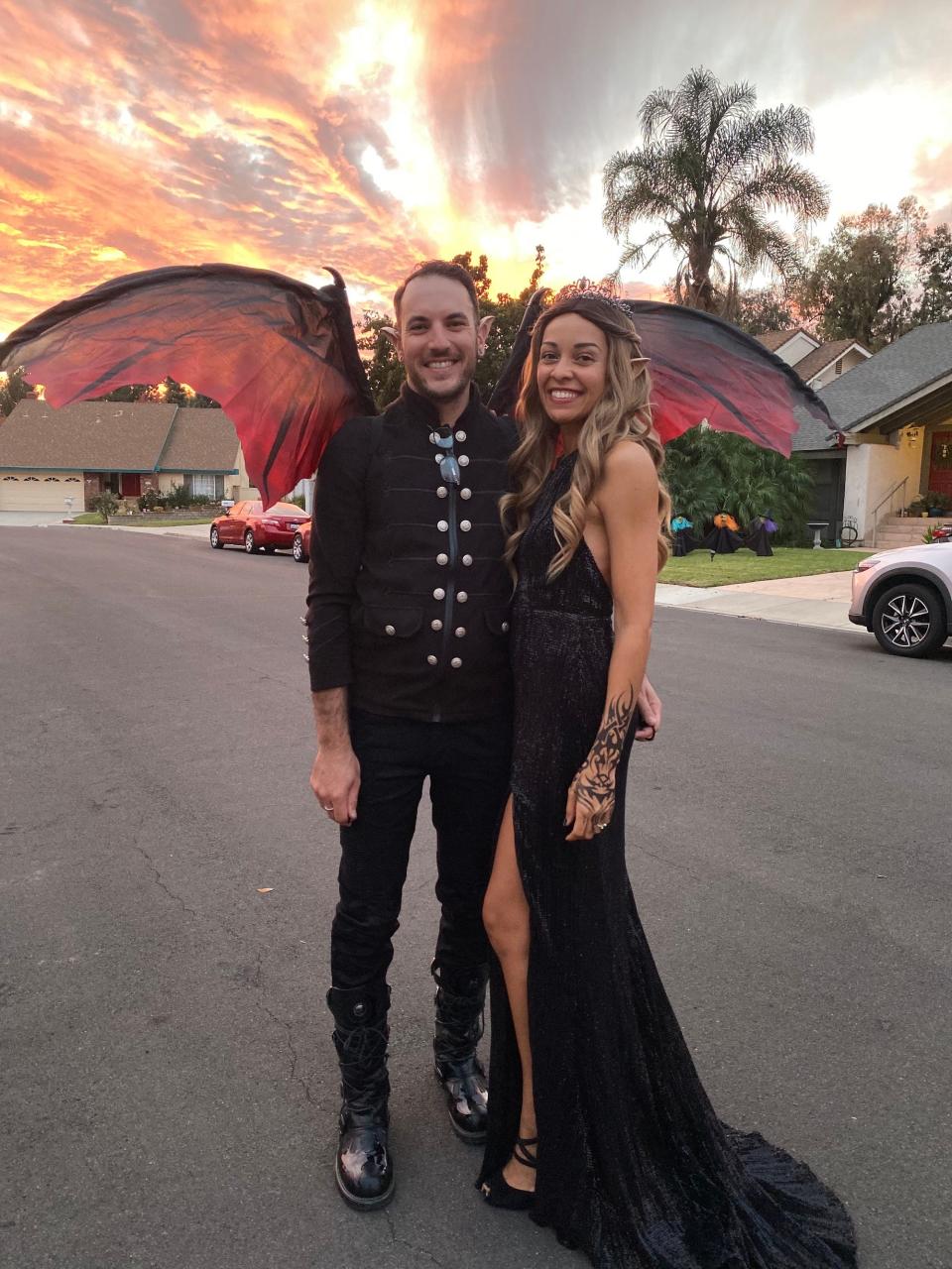 A man in a black suit and red wings stands next to a woman in a black dress.