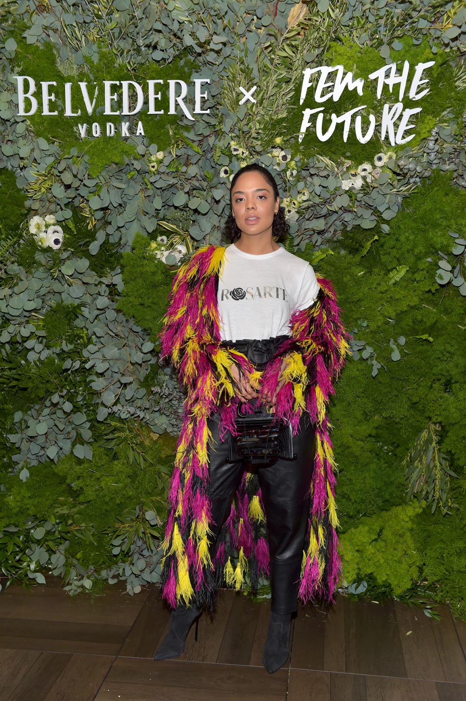 Actor Tessa Thompson attends Belvedere Vodka kick-off "A Beautiful Future" Campaign. She wears black leather trousers, black suede boots, a white t-shirt that says "Rosarte" on it, and a long feathered jacket in black, yellow and fuchsia.