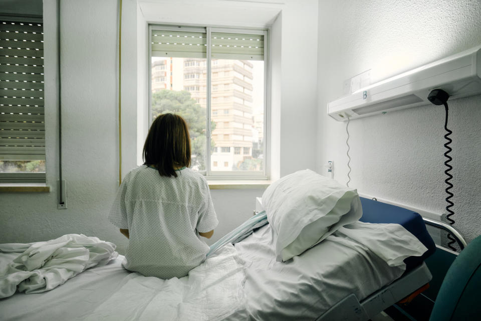 A person in a hospital gown sits on a bed facing a window, looking out at buildings outside