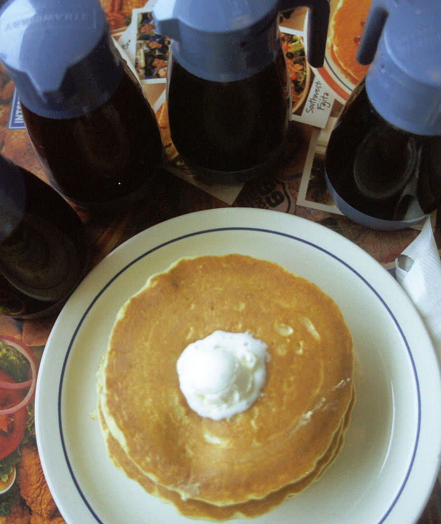 Stack of pancakes topped with butter, surrounded by several syrup bottles. Background shows a colorful, food-themed table mat