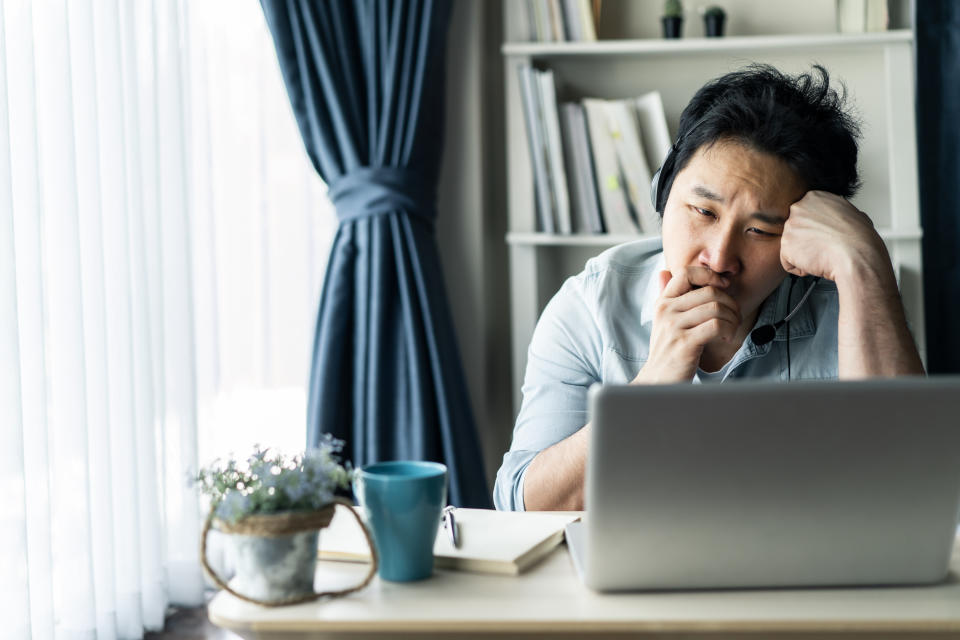 Asian young businessman working from home, man feeling tired, bored and sleepy sitting on table. He looking at computer thinking about job in living room at house, home office due to covid19 pandemic.