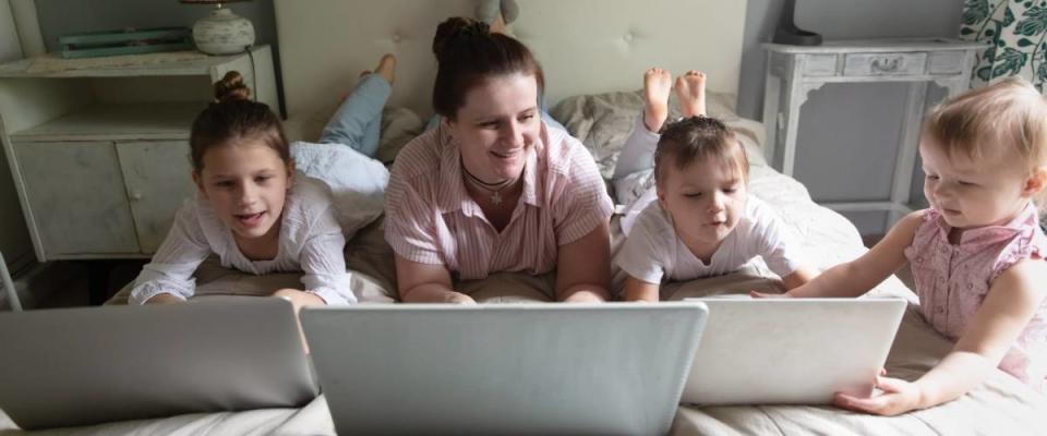 Caucasian Mom and Three siblings children lie on bed with laptops, concept online remote work.