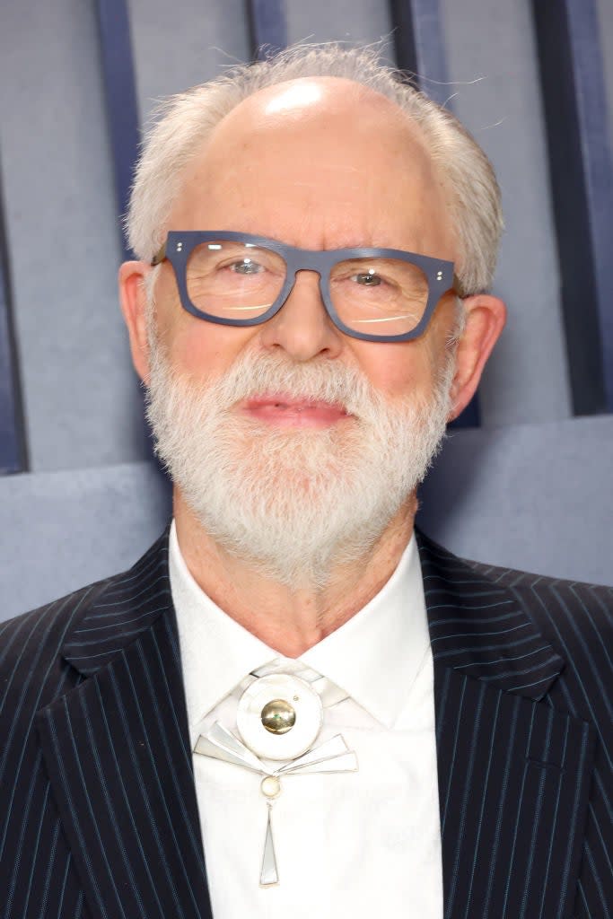 Man with a beard wearing glasses, striped suit, white shirt with a unique circular brooch