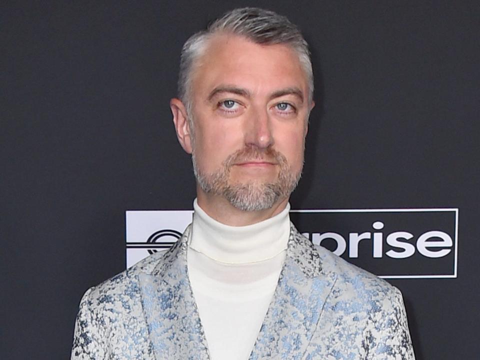 Sean Gunn wearing a light grey jacket and a white turtleneck in front of a black background with an Enterprise Rent-A-Car logo.