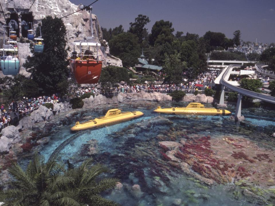 Two large yellow half-underwater submarines in clear water with lots of rocks and plants seen from above. A mountain and cablecars are seen to the left and a steel monorail is seen to the right.