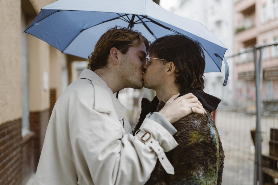 Two people, wearing coats, kiss passionately under an umbrella on a city street