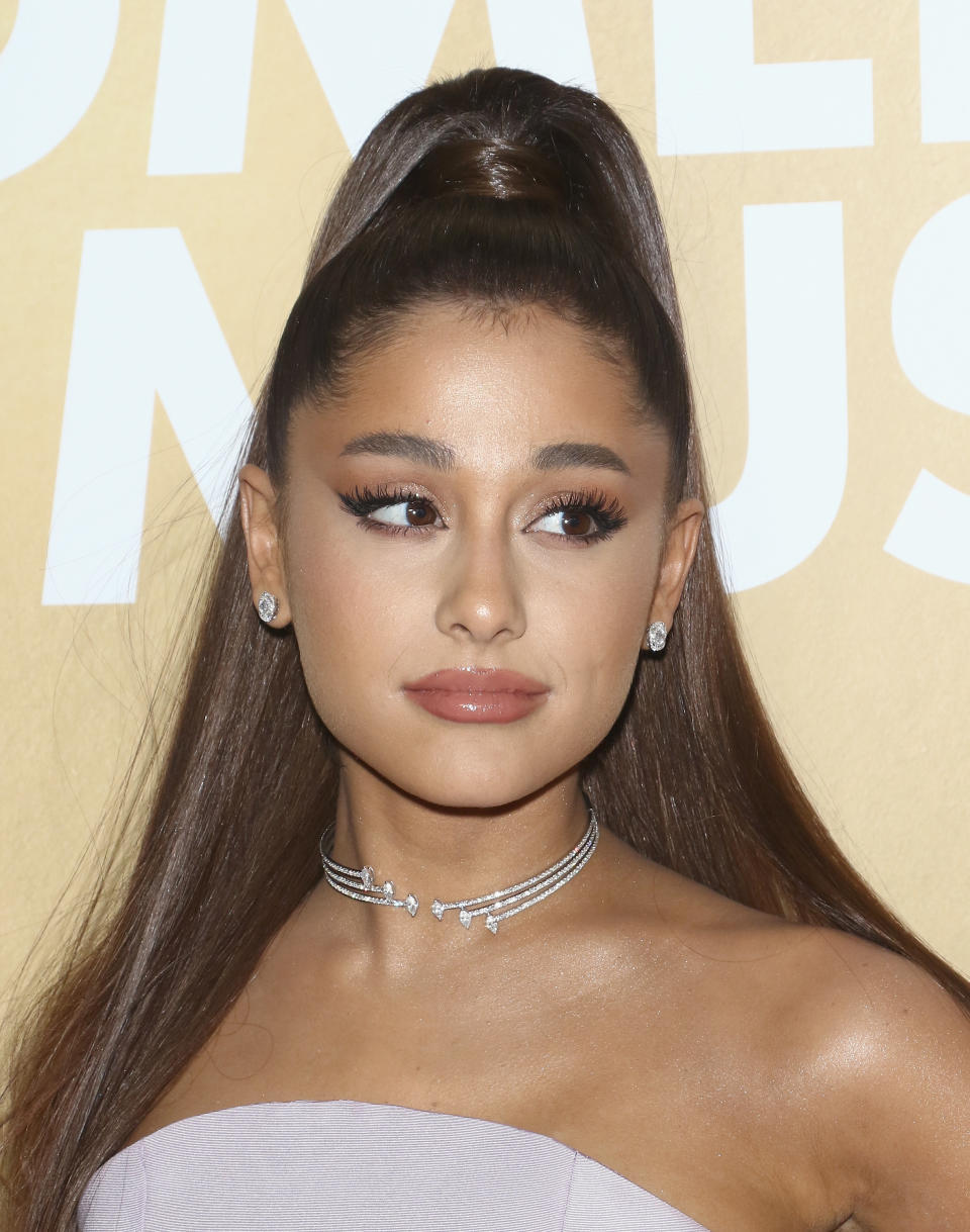 Ariana Grande is wearing a strapless dress and a multi-strand choker necklace with a high ponytail. She stands in front of a backdrop with partially visible text