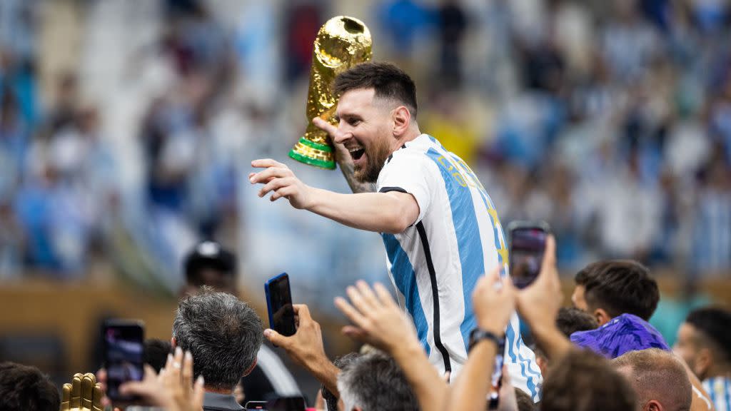 lionel messi holding the world cup trophy and smiling as he is carried by teammates