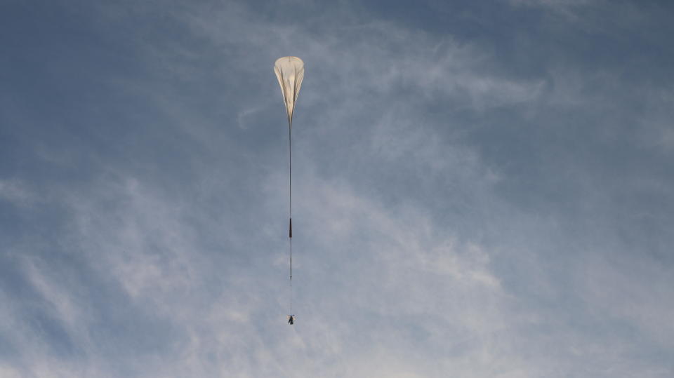 a large white balloon carries a dark, cylindrical payload through a blue sky.