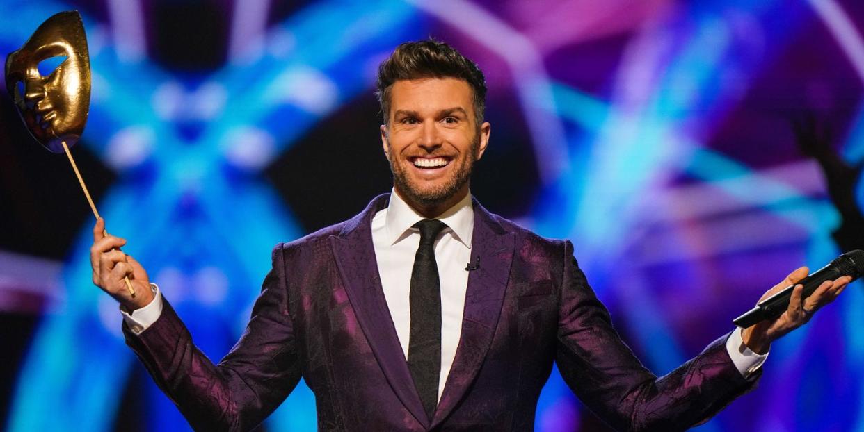 the masked dancer host joel dommett on stage, holding a gold theatre style face mask on a stick in his right hand and his microphone in his left hand