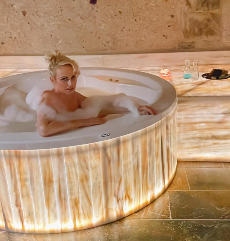 Rebel Wilson sits nude in a bathtub, covered in bubbles.