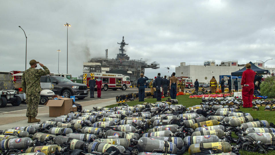 Sailors stand at attention and salute the national ensign during morning colors behind the amphibious assault ship USS Bonhomme Richard (LHD 6) at Naval Base in San Diego, Calif. on Tuesday, July 14, 2020, as the ship endures an ongoing fire. The fight to save a burning Navy warship docked in San Diego has entered a third day with indications of improvement. A Navy statement says there's been significant progress and much less smoke is being emitted from the USS Bonhomme Richard on Tuesday. (Mass Communications Specialist 3rd Class Jason Waite/U.S. Navy via AP)