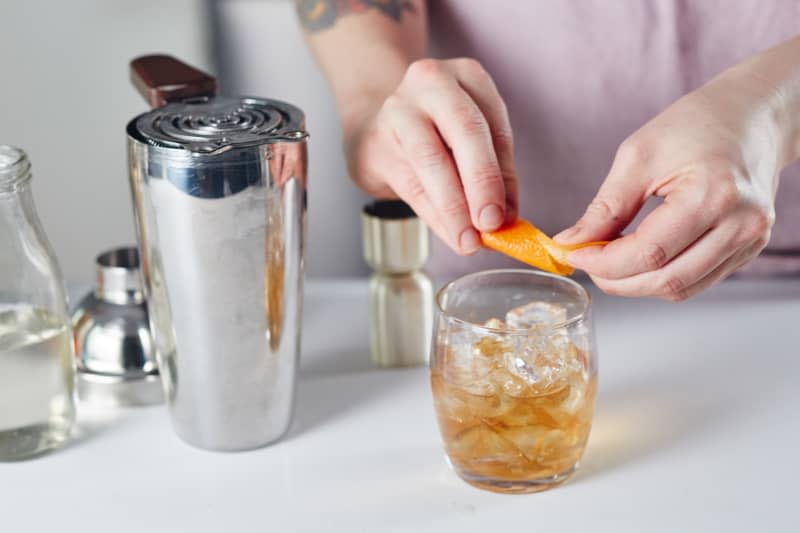 Someone uses an orange peel as a garnish, with bartending tools in the background