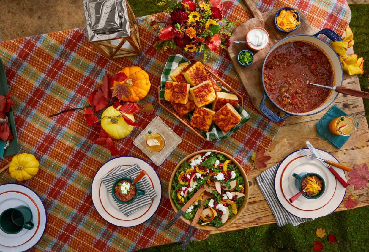 a table with chili and cornbread and a green salad and cups of the chili and some drinks and there are pumpkins decorating the table too