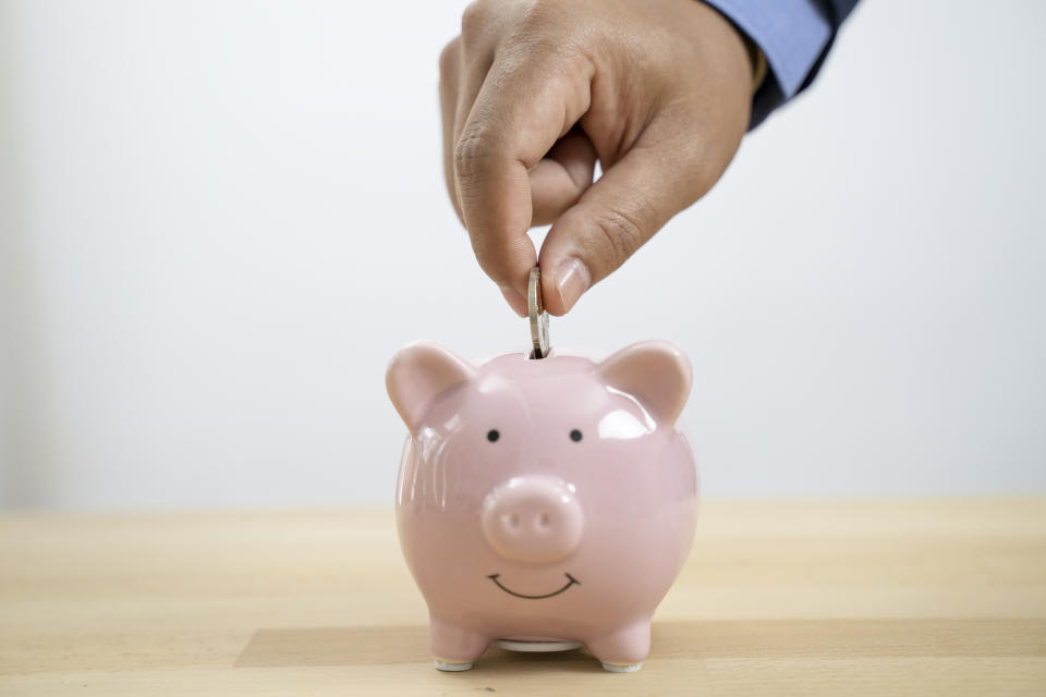 A hand is placing a coin into a pink piggy bank with a smiling face, symbolizing saving money
