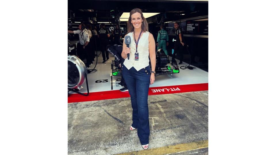 Lee McKenzie holding a microphone in front of an F1 garage