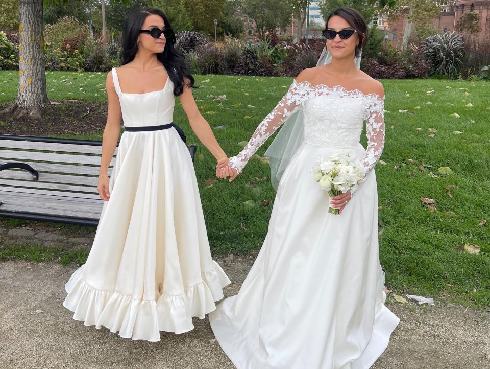 Two women in wedding dresses and sunglasses hold hands.