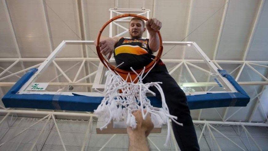 Ben sat on top of an indoor basketball hoop wearing a black vest. The photographer is looking up at him through the hoop net