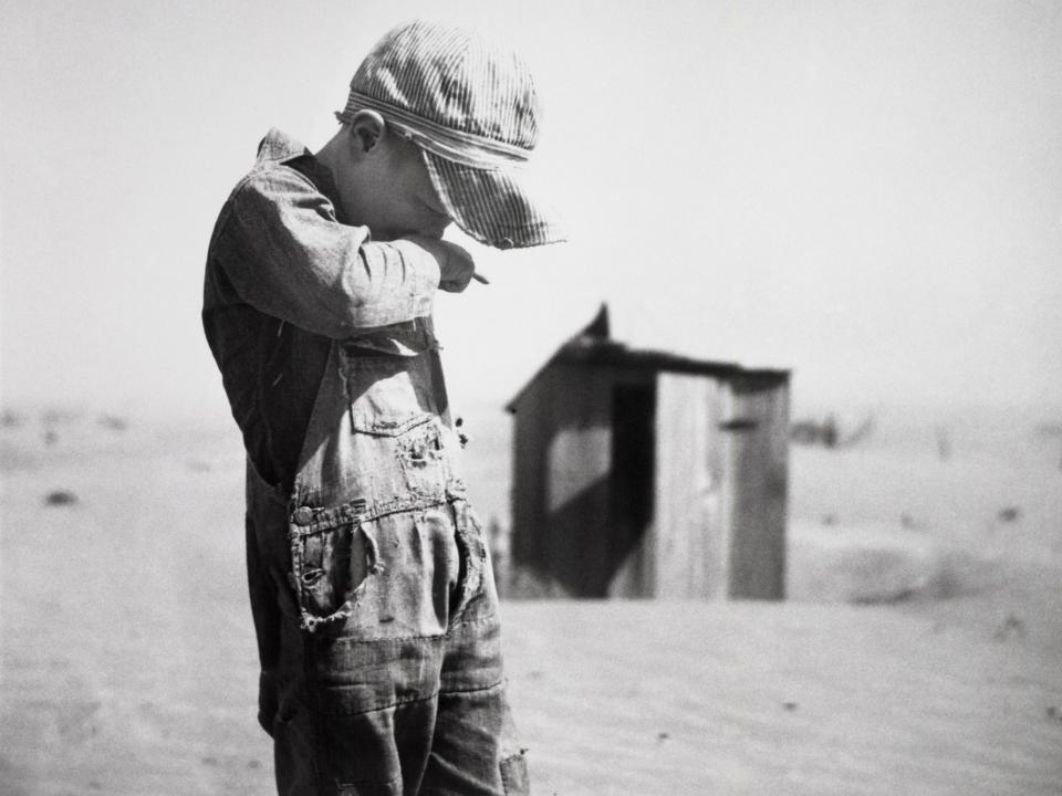 A young boy covers his nose and mouth against brown sand in the Dust Bowl, circa 1930.