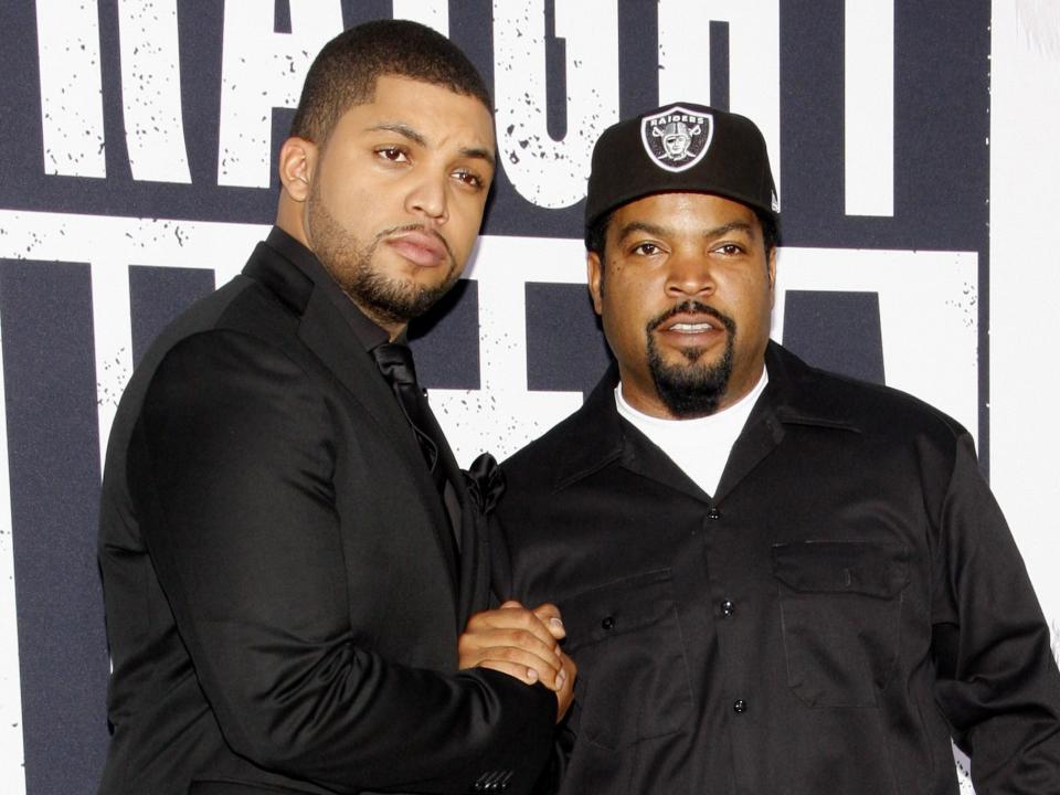 A young O'Shea with facial hair and a black shirt next to an older Ice Cube with facial hair, a hat, and a black shirt.