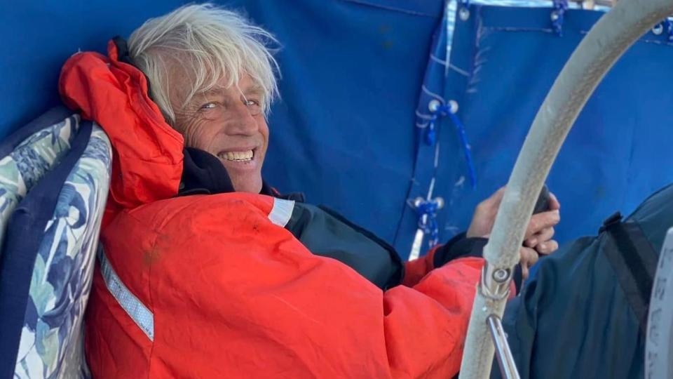 A smiling Brett Clibbery huddled on the deck of his yacht the Theros. He is wearing a red waterproof sailing jacket.