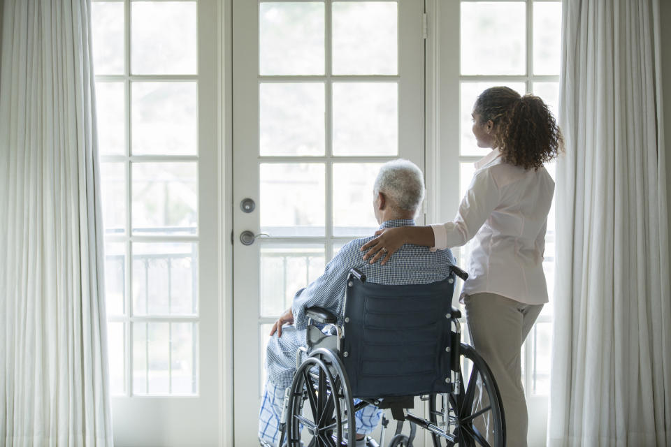 An elderly person in a wheelchair and a younger person standing beside them look out a window, sharing a moment of closeness and care