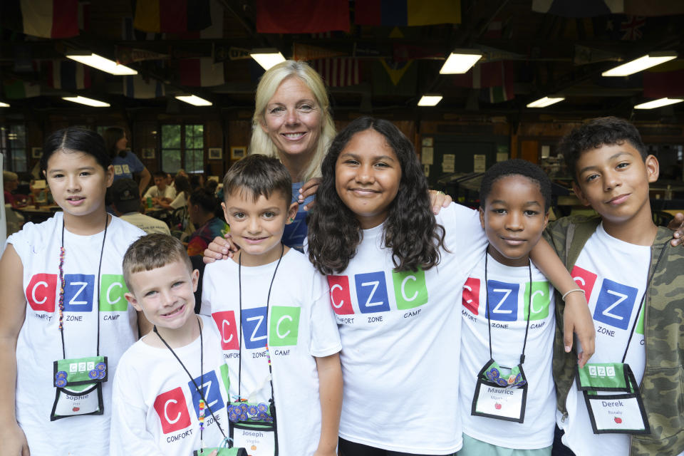 Six children of mainly middle school age, wearing lanyards that show their names and wearing T-shirts printed with CZC, Comfort Zone Camp, pose with a woman in a large, wood-paneled community hall. 