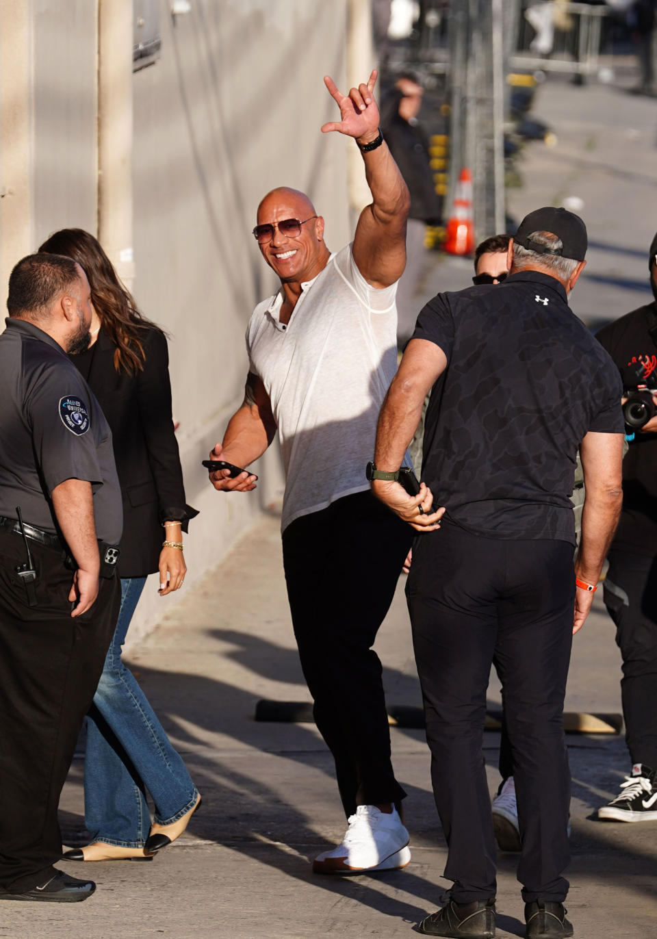 Dwayne Johnson waves while walking, flanked by security, in a casual v-neck and jeans