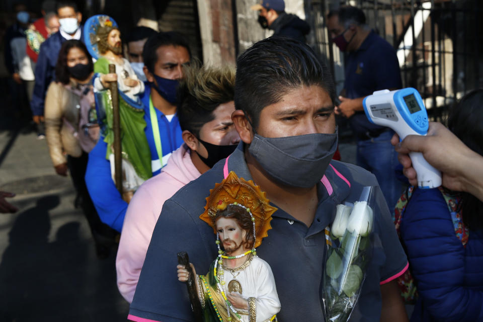 A man holding a statue of Saint Jude and wearing a protective face mask amid the new coronavirus, stands still as his temperature is measured before entering the San Hipolito Catholic church, during the annual pilgrimage honoring Jude, the patron saint of lost causes, in Mexico City, Wednesday, Oct. 28, 2020. Thousands of Mexicans did not miss this year to mark St. Jude's feast day, but the pandemic caused Masses to be canceled and the rivers of people of other years were replaced by orderly lines of masked worshipers waiting their turn for a blessing. (AP Photo/Marco Ugarte)