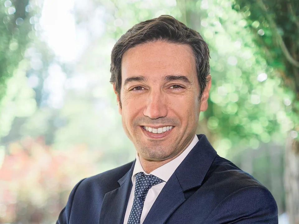 A headshot of Raul Vargas smiling while standing in a brightly lit garden and wearing a suit and tie .