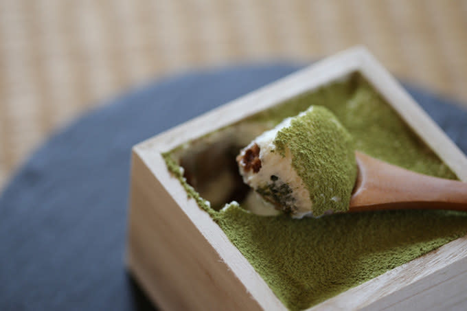 Tiny wooden spoon scooping up a bit of matcha tiramisu from a wooden box.