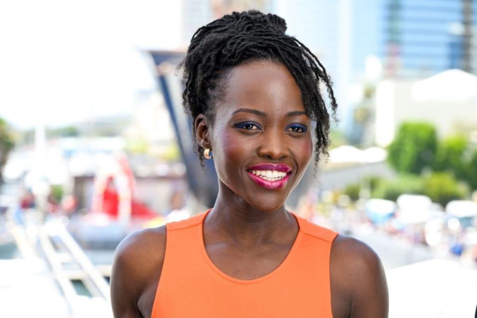 Lupita Nyong'o smiles while wearing a sleeveless, fitted dress