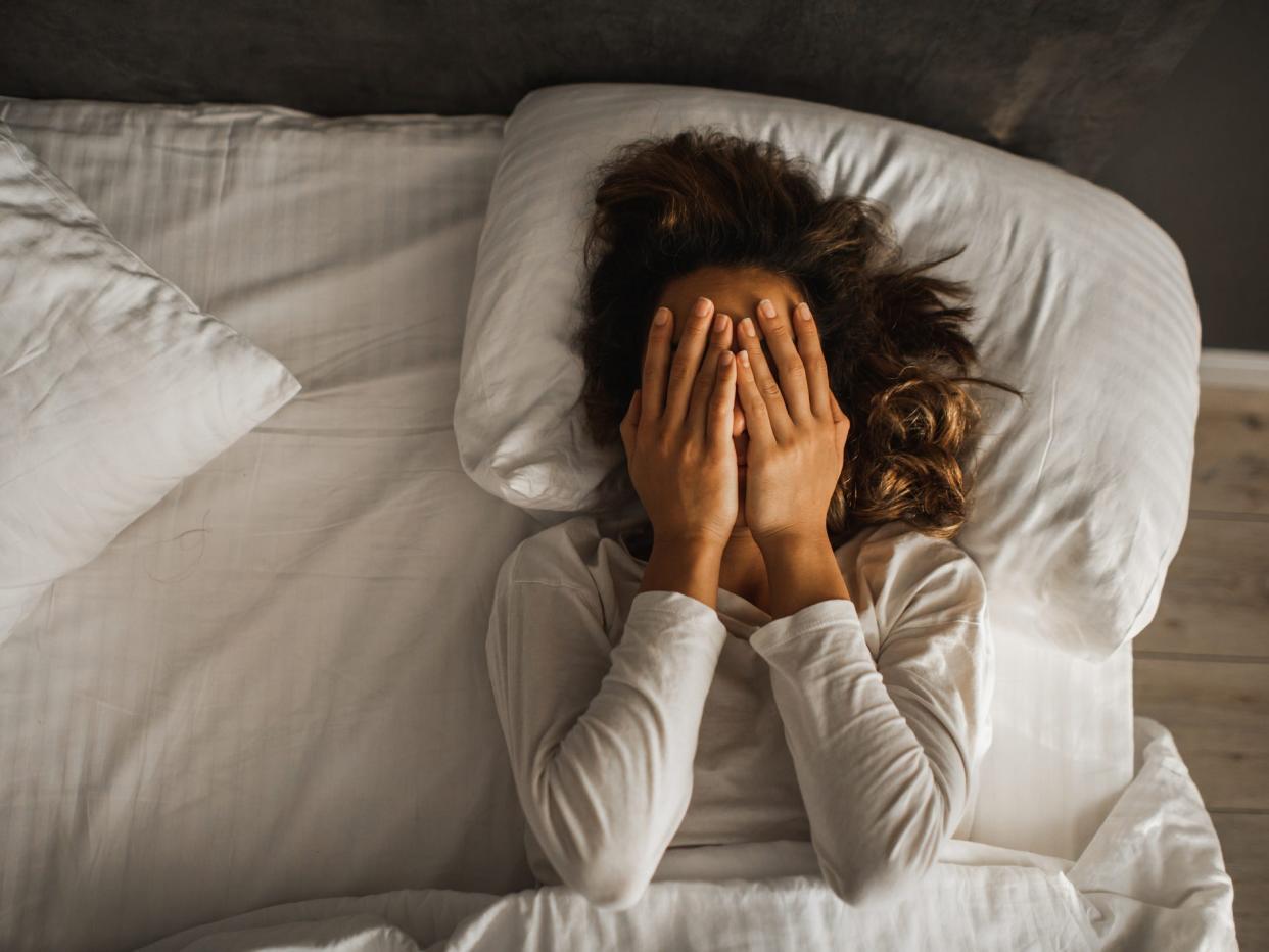 Photo from above of a woman with light brown skin lying in a bed with white striped sheets. She covers her face with her hands. Her light brown hair poofs out around her head on the pillow and flows over her left shoulder. She wears a long sleeve white shirt. To her left, you can see a wooden floor stained white and a wall painted a dark color.