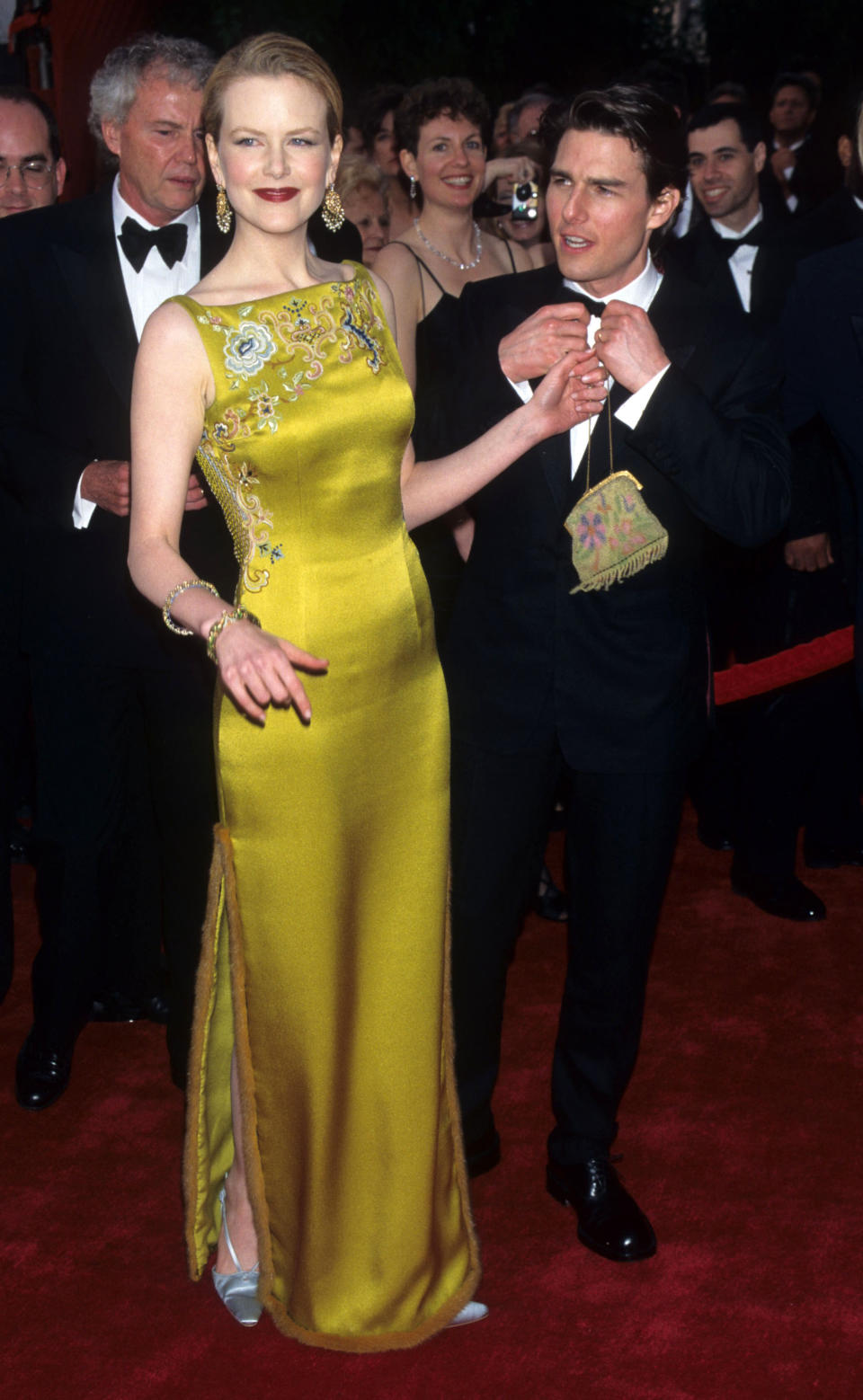 Nicole Kidman stands on a red carpet in a green/yellow floor-length dress, she's handing her bag to Tom Cruise who stands behind her in a tux