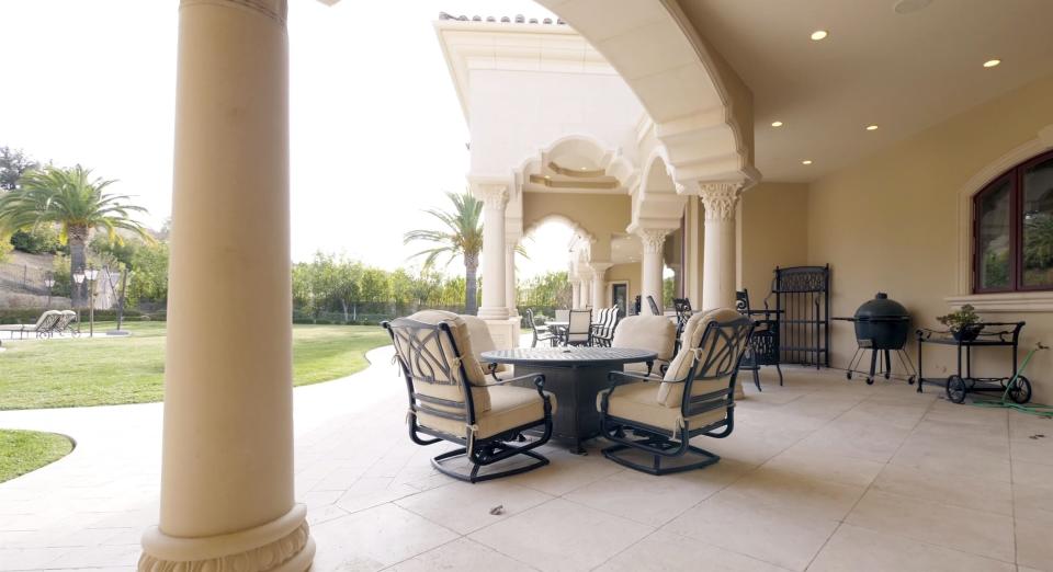 A curved patio holds a circular table with chairs situated around it, as well as a grill and a rolling cart with a plant on it