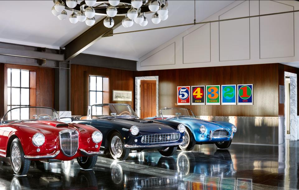 A 1955 Lancia Aurelia B24S Spider America, A 1960 Ferrari 250 GT Cabriolet Series II, and a 1964 Shelby Cobra 289 lined up in the main room. On back wall, a set of Robert Indiana numbers.