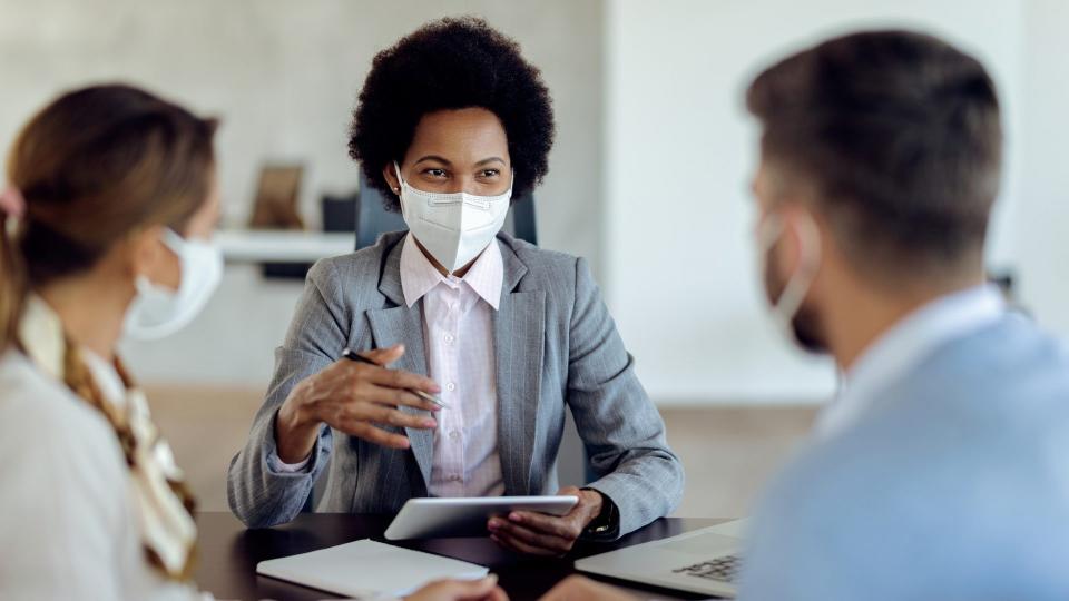 Happy black bank manager with face mask using touchpad while talking to her clients on a meeting in the office.