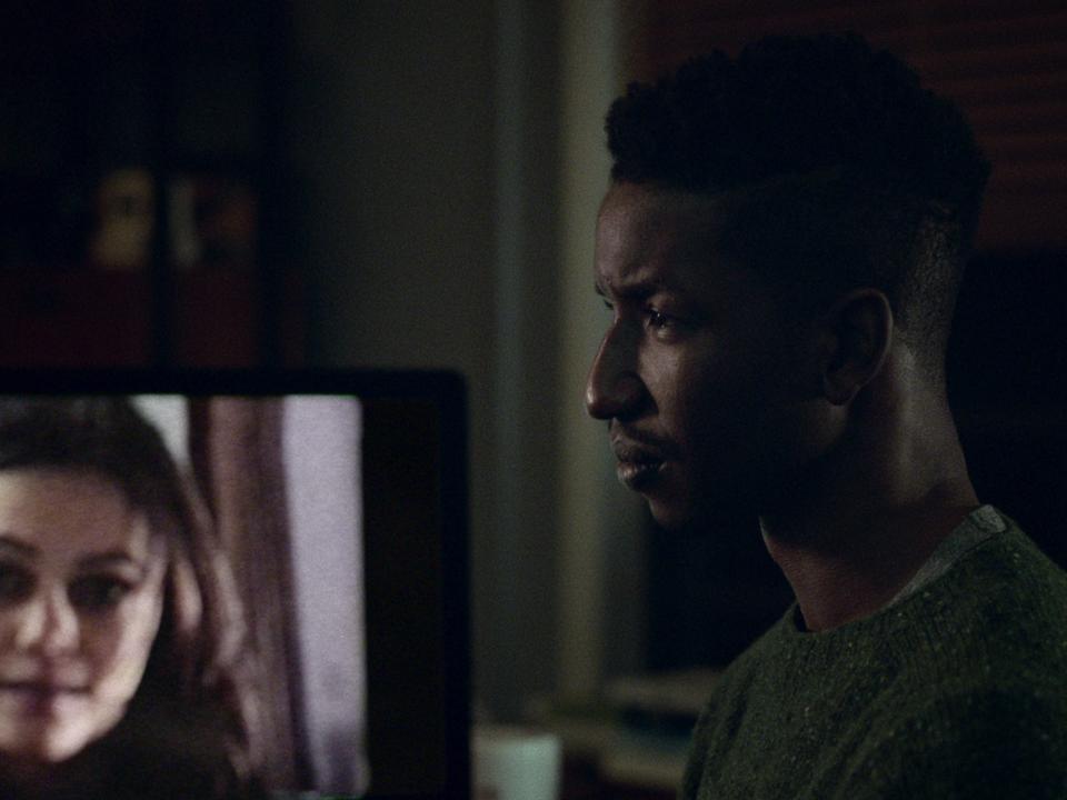 Mamoudou Athie standing in front of screen with woman's face on it in "Archive 81"