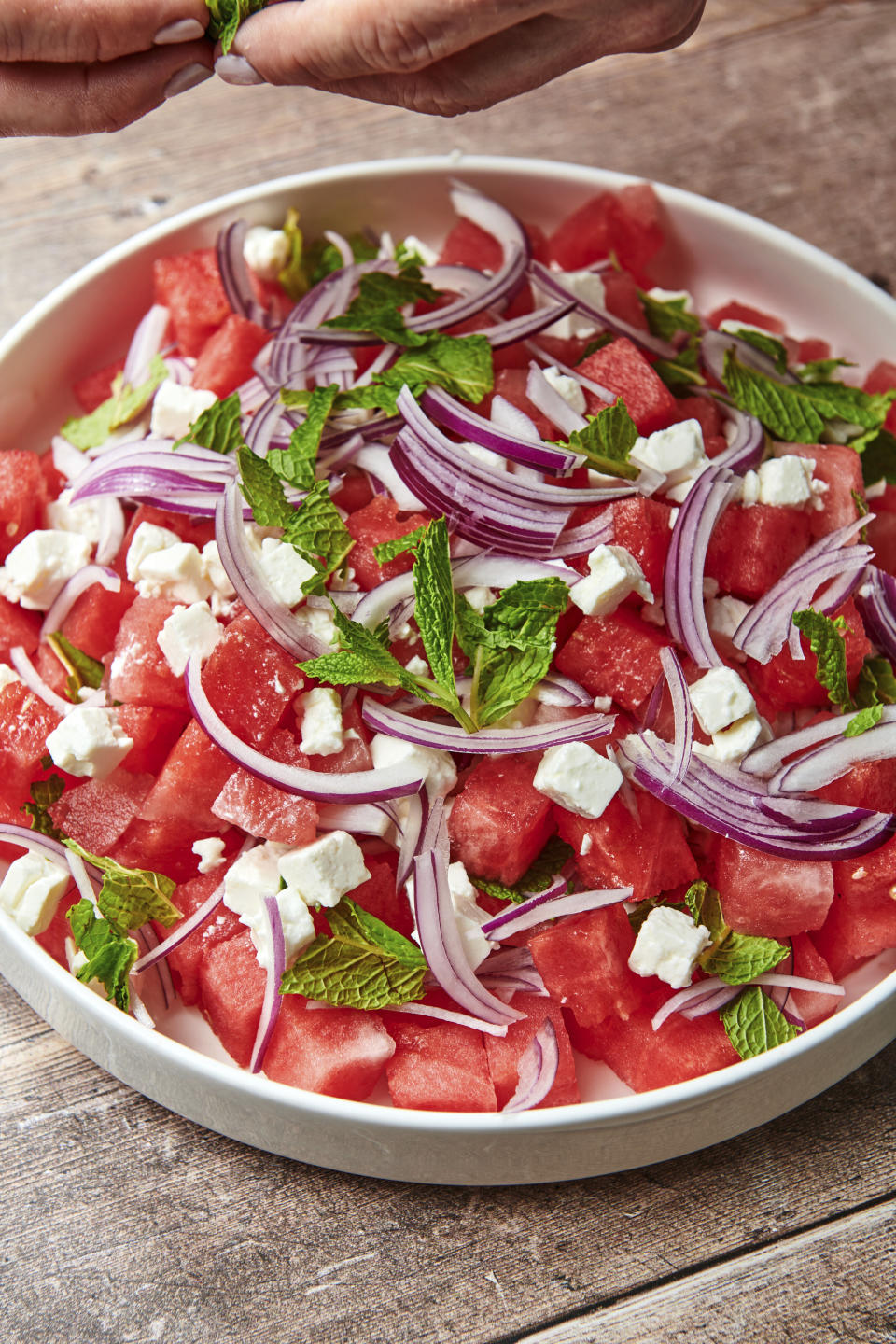 This image shows a recipe for watermelon feta salad topped with thinly sliced red onion and mint leaves. (Cheyenne Cohen via AP)