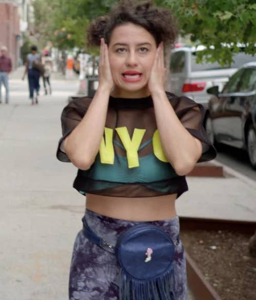 Ilana Glazer holds her face with both hands, wearing a black sheer crop top with "NYC" letters over a green tank and a blue floral skirt