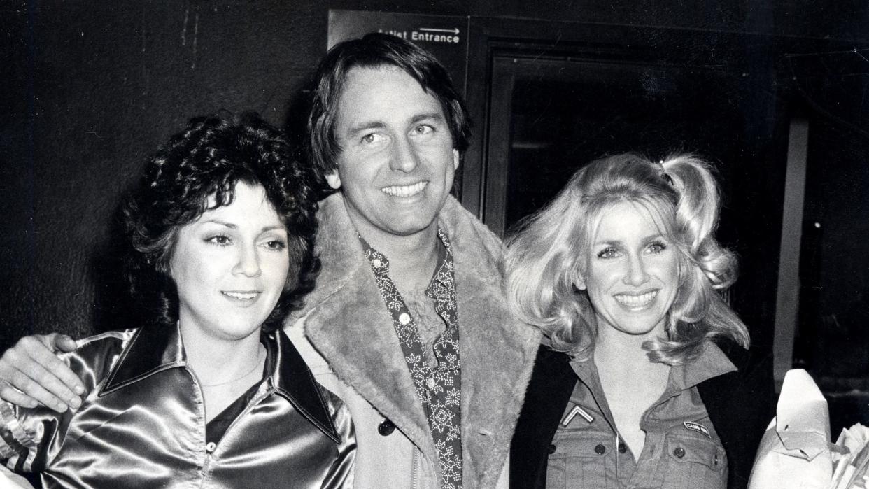 joyce dewitt, john ritter, and suzanne somers smile and pose for photos while standing outside a building, ritter has his arms around both women as they each hold supplies in one arm