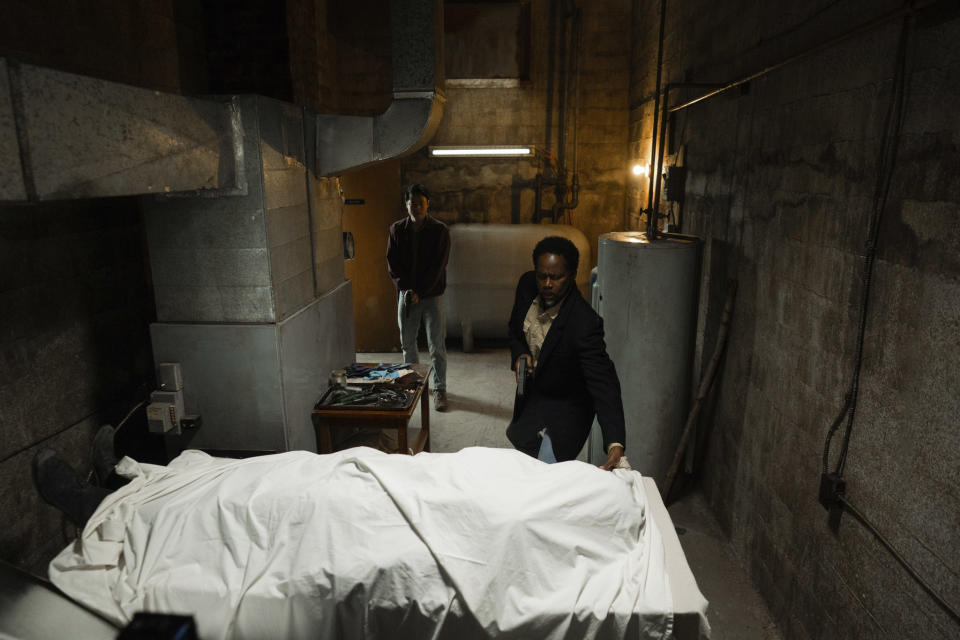 Two men approach a dead body under a sheet in an empty room