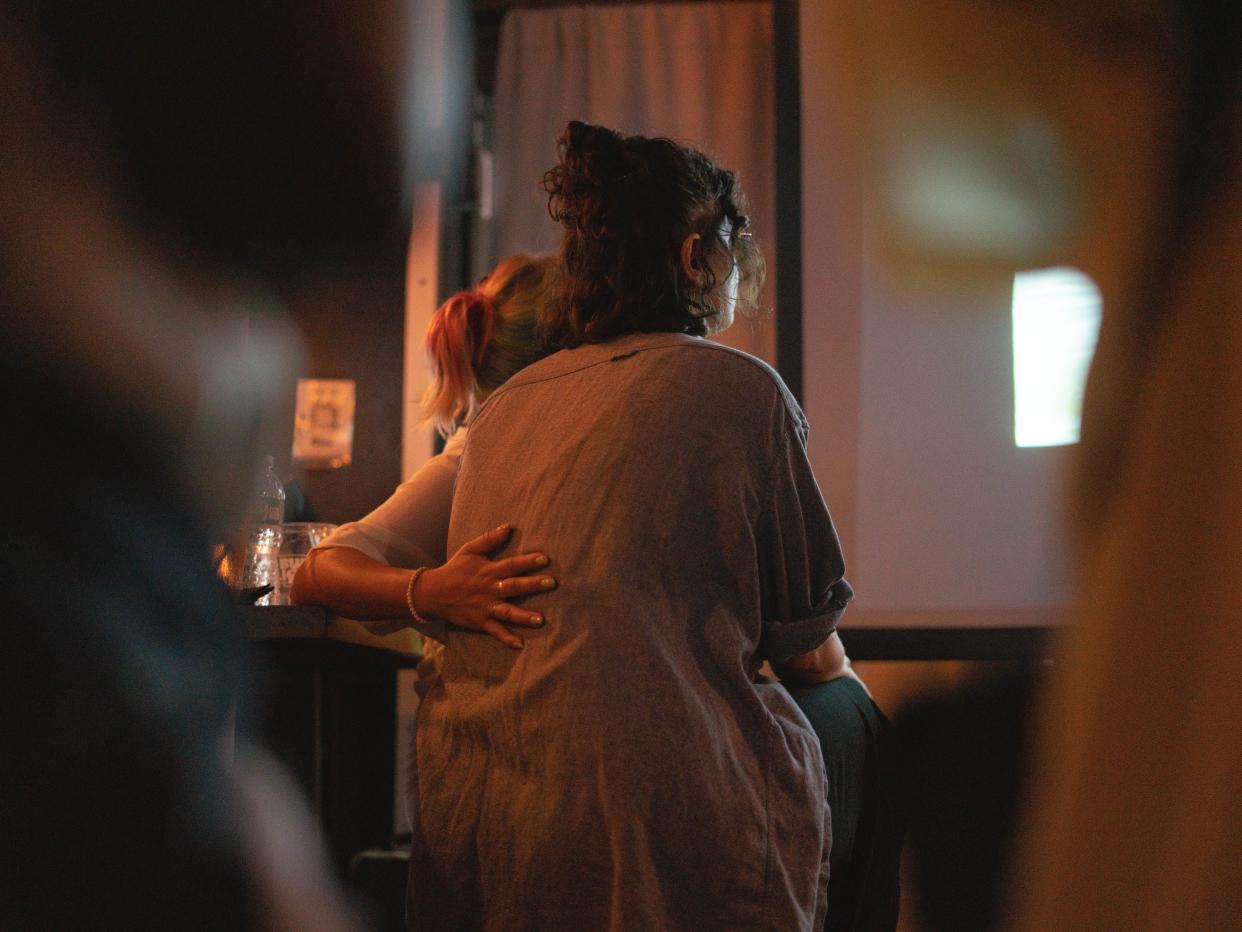 viva, a person with colored hair in a ponytail, puts their hand around violet augustine, as she watches moving isa, a documentary about her daughter