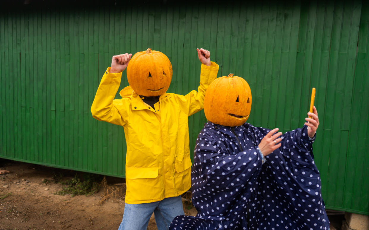 Best couples Halloween costumes in 2023, couple wearing orange pumpkins on heads in yellow rain jacket and jeans and navy blue polka dot poncho taking selfie against green background