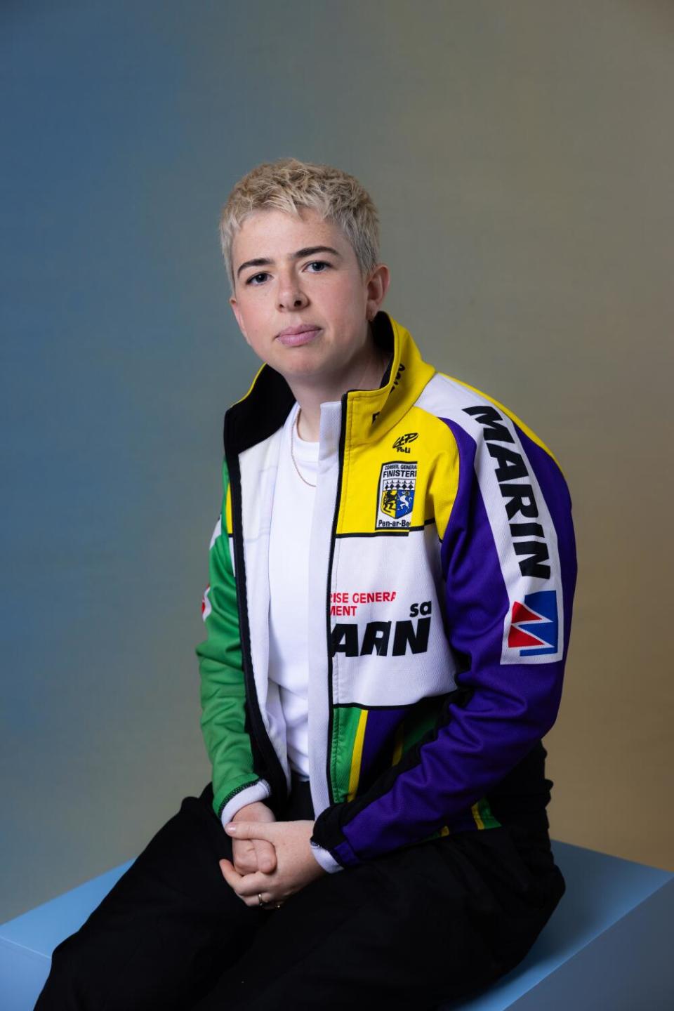 Director Molly Manning Walker wears a blue, green, white and black open jacket while sitting on a cube.