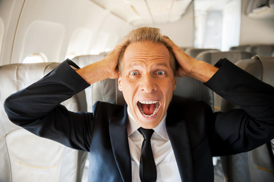 Shocked mature businessman touching his head with hands and looking at camera while sitting at his seat in airplane