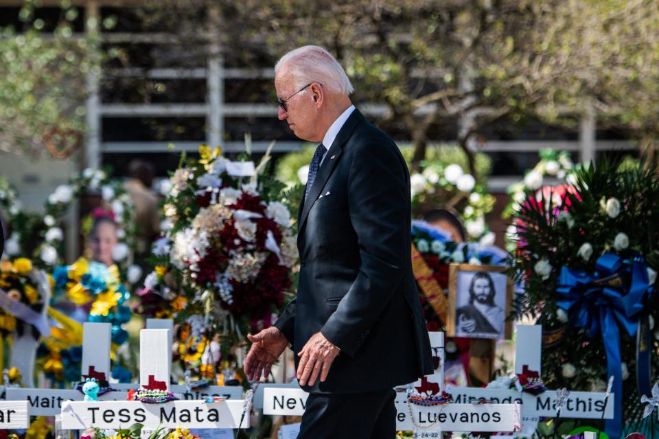 President Biden, in a dark suit with sunglasses, stands among a display of flowers, white crosses with names and a small framed picture of Jesus.